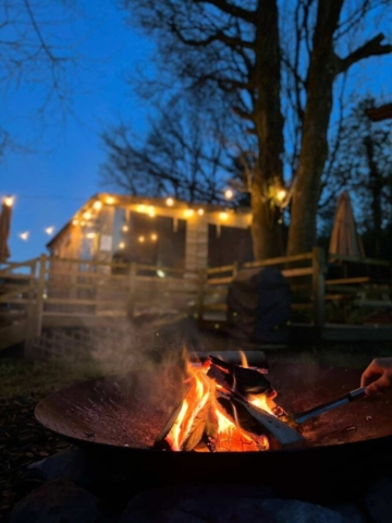 A person cooks over an open fire in front of a cozy cabin, surrounded by nature and the warmth of the flames.