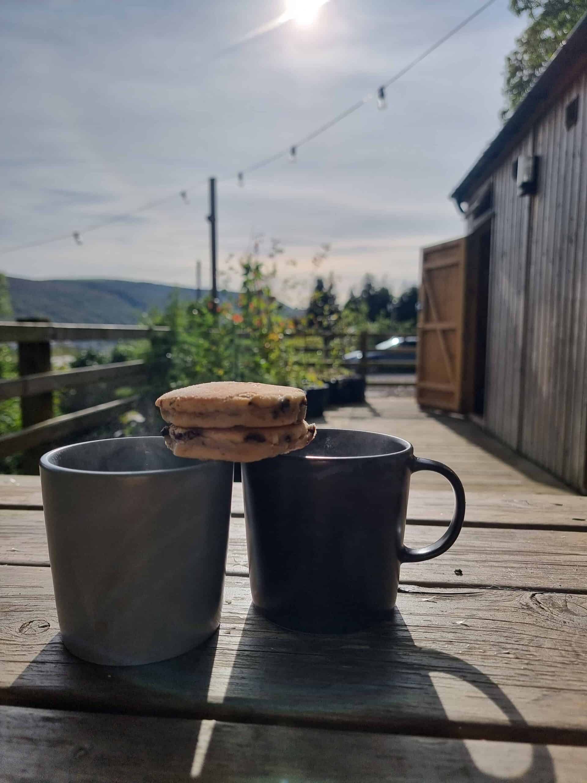 Mugs of hot drinks with welsh cakes sat on a table against a back drop of a clear sky and hills