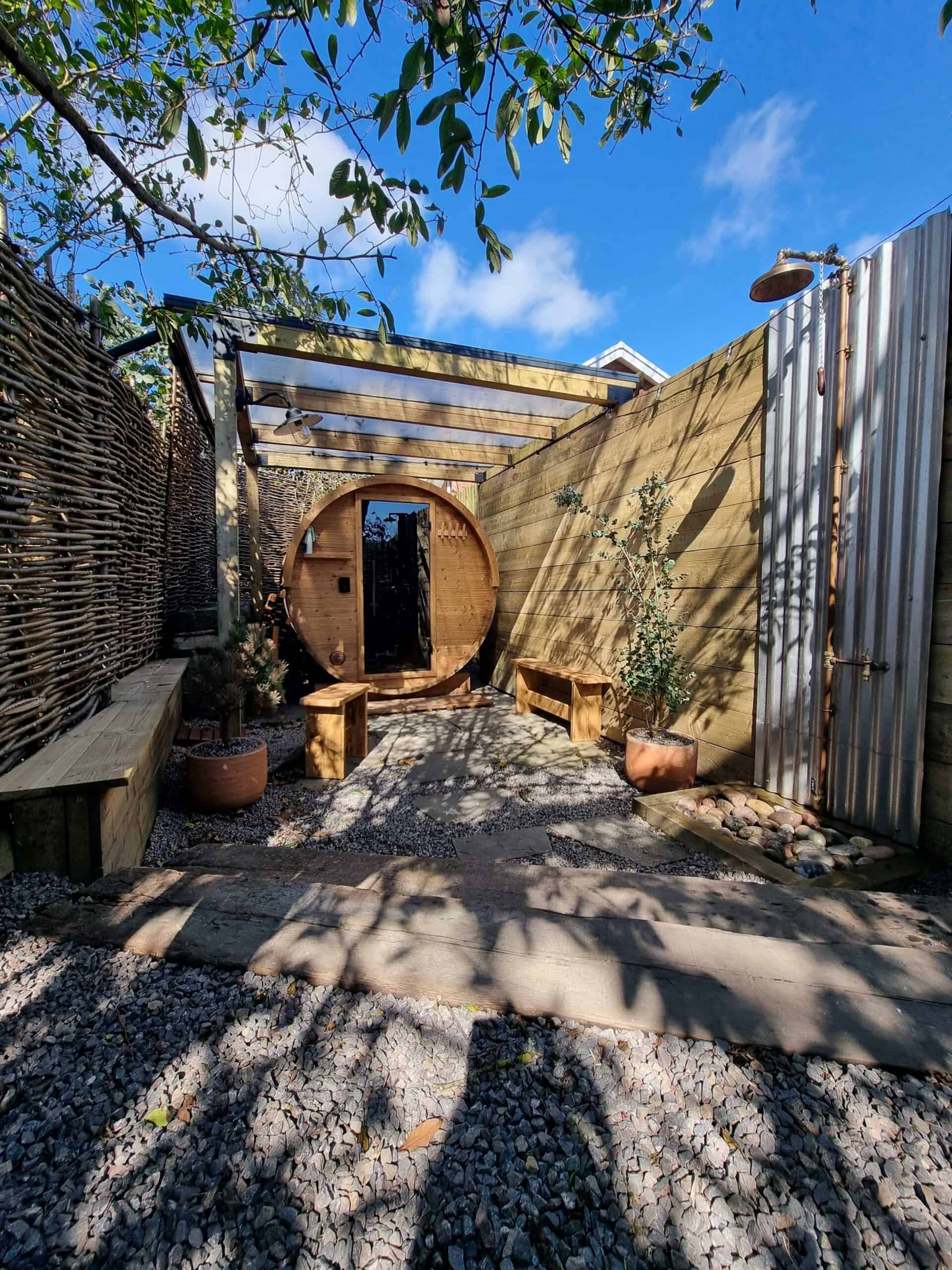 A round outdoor wooden sauna under the trees with an outdoor shower and bench seats