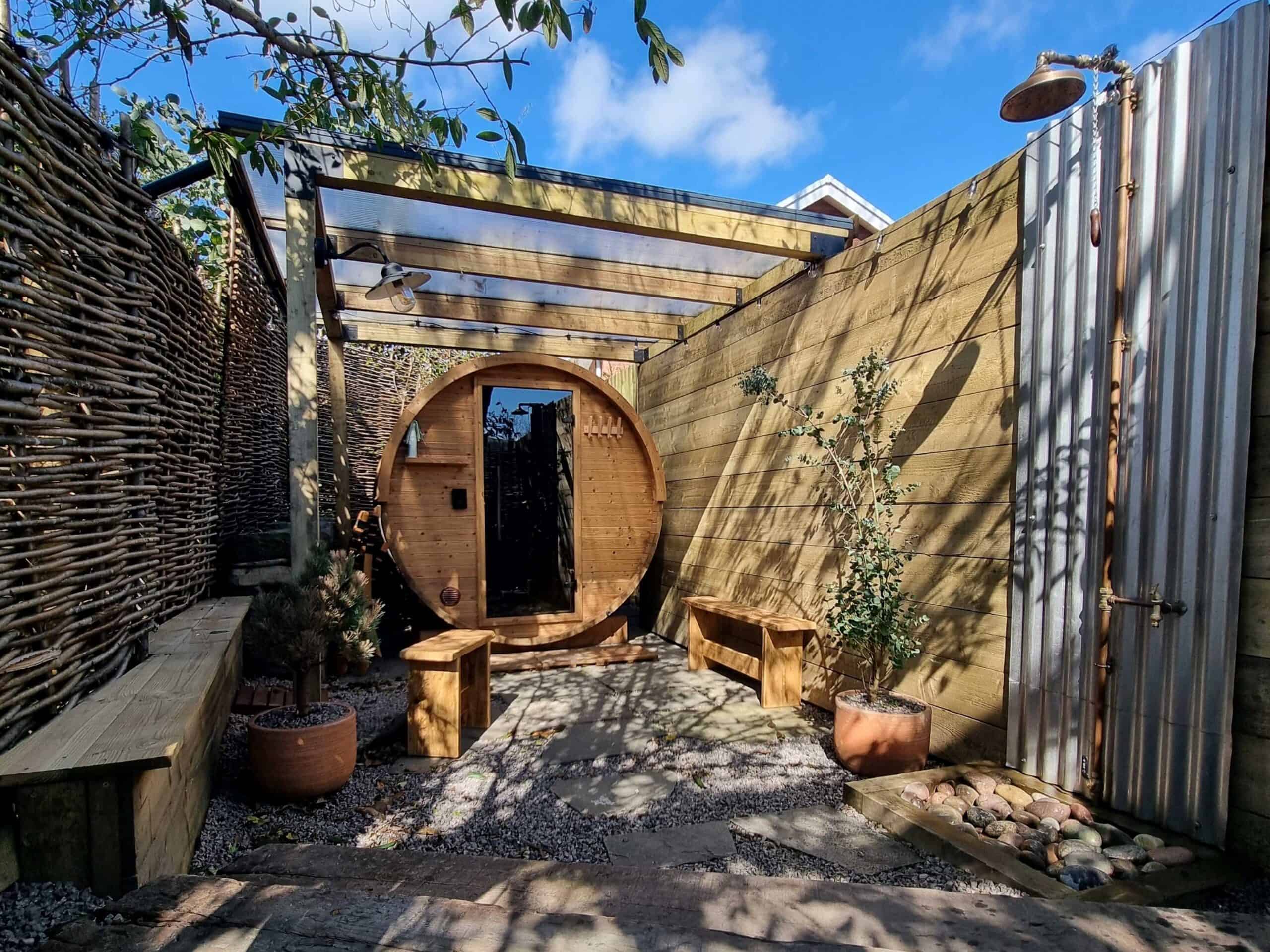 A round outdoor wooden sauna under the trees with an outdoor shower and bench seats